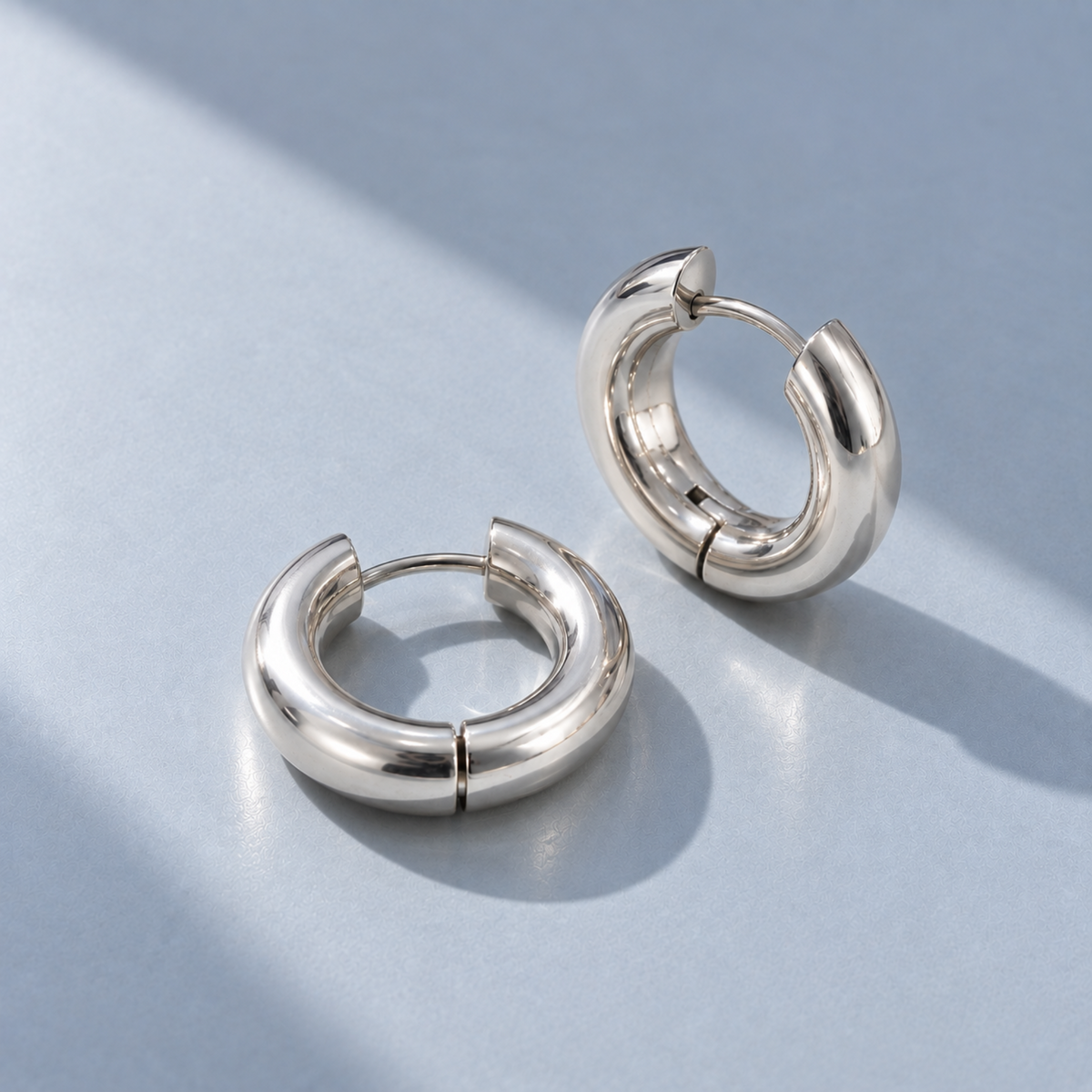 A pair of polished silver hoop earrings with a thick, rounded modern design, displayed on a light blue-gray surface with soft shadows.