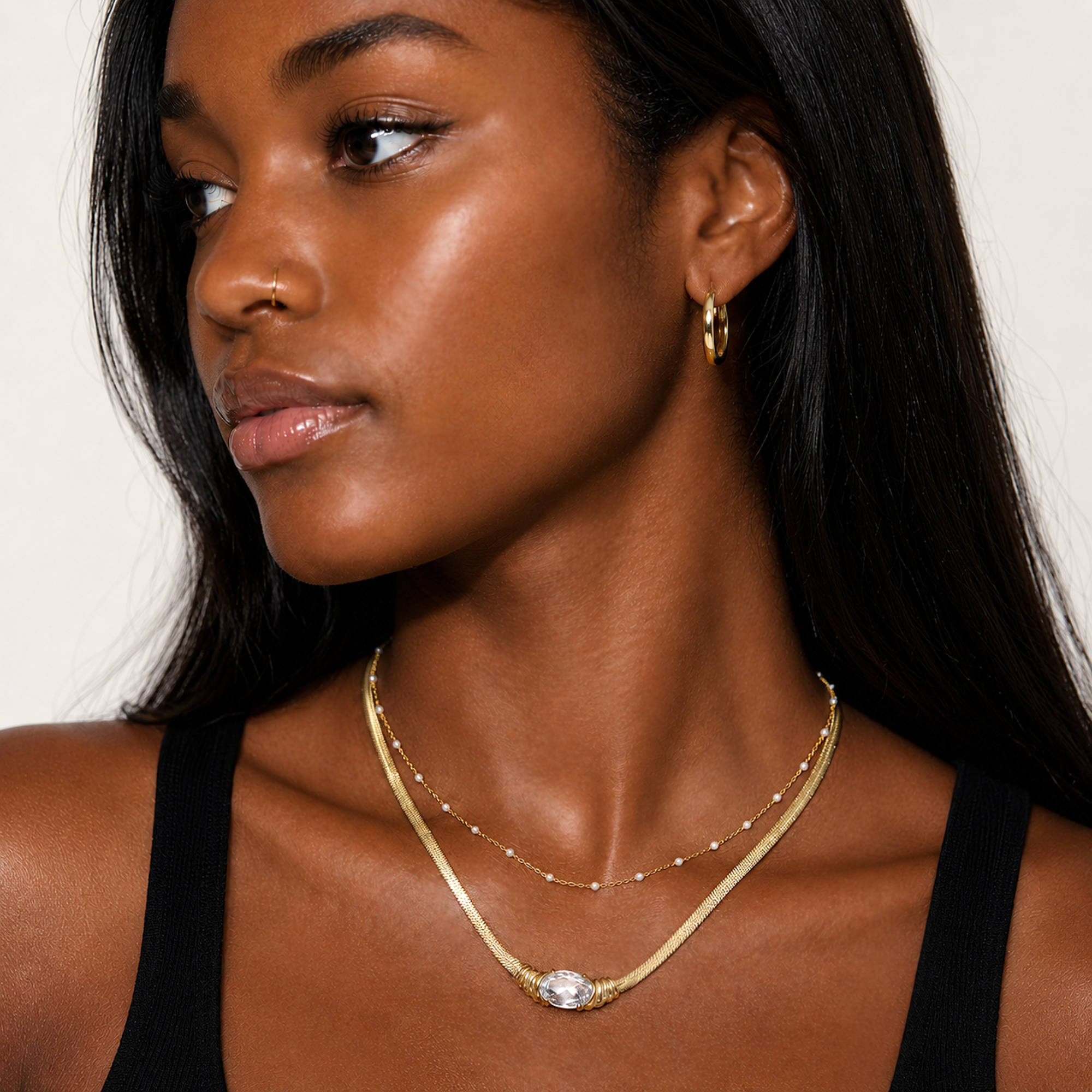 Woman wearing a dainty white beaded gold chain gold necklace stacked with a white pendant snake chain necklace and gold hoop earrings on a neutral background